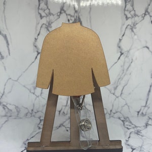 Puede incluir: Un caballete de madera marrón con un recorte de madera marrón de un suéter colgando de él. El suéter tiene cuello alto y mangas largas. Un portagafete de plástico transparente con un clip plateado está unido al suéter.