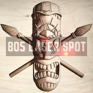 May include: A wooden tiki mask with a carved face and crossed spears behind it. The mask has a large mouth with teeth and a detailed design on the forehead. The text "805 LASER SPOT" is visible below the mask.