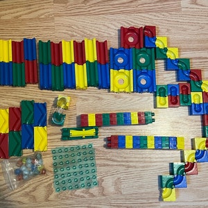 May include: A colorful marble run set with various plastic components in red, blue, green, and yellow. The set includes straight tracks, curved pieces, and a collection of marbles. The pieces are arranged on a wooden surface.