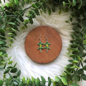 May include: A pair of beaded lizard earrings. The earrings feature a green and yellow color scheme, with a lizard design. The earrings are displayed on a wooden disc, surrounded by greenery and a white, fluffy surface.