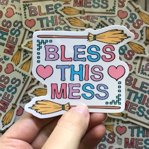 May include: A white sticker with a colorful design that says "Bless This Mess" with two pink hearts and two broomsticks. The sticker has a pixelated border.