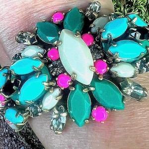 Op de afbeelding: Een gouden armband met turquoise, groene en witte stenen. De stenen zijn in de vorm van een bloem gerangschikt.
