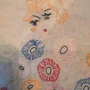 May include: Embroidered artwork featuring a woman's face with orange hair and red lips. The woman is surrounded by colorful embroidered flowers in shades of blue, red, yellow, and white. The artwork is on a light blue background.