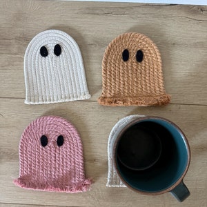 May include: Four handmade crocheted ghost-shaped coasters in white, brown, pink, and beige. The coasters are on a wood surface. A dark green mug is sitting on top of one of the coasters.