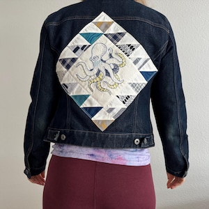 May include: A dark blue denim jacket with a white, blue, and yellow patchwork design on the back. The design features an embroidered octopus.