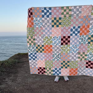 May include: A colorful patchwork quilt with a checkerboard pattern. The quilt features squares in various colors, including blue, orange, green, and pink. Some squares have animal prints. The quilt is held up against a backdrop of the ocean and sky.