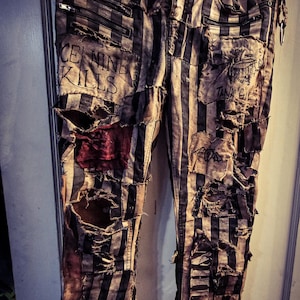 Op de afbeelding: Verouderde zwart-wit gestreepte broek met meerdere scheuren en patches. De broek heeft ritsen en de tekst "ICE NINE KILLS" en "TANK GIRL". De broek hangt aan een metalen hanger.