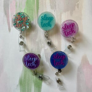 May include: Five colorful badge reels with the text "Sleep Tech". The reels are round and feature glitter and various colors, including teal, purple, and blue. Each reel has a clear plastic clip and a retractable cord.