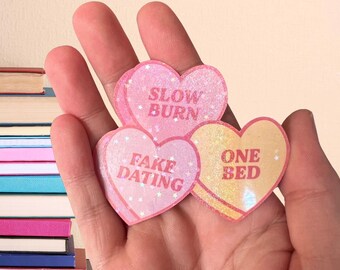 May include: Three pink heart-shaped stickers with glitter. The stickers have the following text: "Slow Burn", "Fake Dating", and "One Bed".
