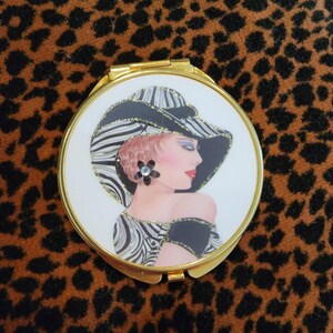 Peut inclure: Un miroir compact doré avec un fond blanc et une illustration en noir et blanc d'une femme portant un chapeau. La femme a les cheveux blonds et porte une robe à rayures noires et blanches.