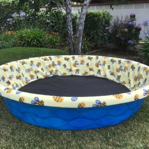 May include: A round, blue pet bed with a yellow and black bee-themed fabric lining the rim. The bed has a black interior and is set on a grassy lawn. The bed's exterior is a textured blue plastic.