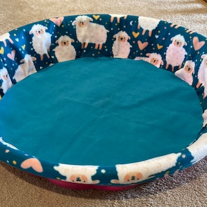 May include: A round pet bed with a teal interior and a patterned exterior. The exterior features a dark teal background with white sheep, pink hearts, and yellow accents. The bed has a pink base.