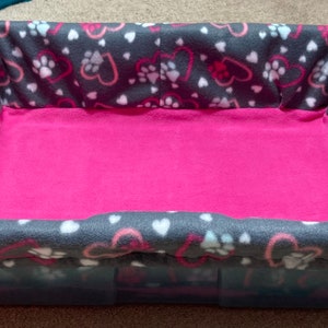 May include: A gray and pink pet bed with a heart and paw print design. The bed is made of soft fleece and is perfect for small dogs or cats.