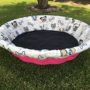 May include: A pink and white dog bed with a black, removable liner. The bed is decorated with a pattern of dogs and bones.