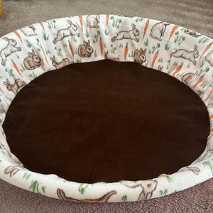 May include: A round pet bed with a brown interior and a white exterior. The exterior features a repeating pattern of brown rabbits, orange carrots, and green foliage. The bed is designed for small pets.