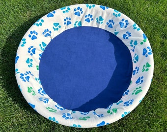 Peut inclure: Lit pour animaux de compagnie rond avec un intérieur bleu et un bord blanc. Le bord est orné d'empreintes de pattes bleues et vertes. Le lit est conçu pour les animaux de compagnie.
