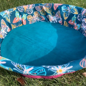 May include: A teal blue and pink pet bed with a butterfly print. The bed is round and has a soft, plush lining.