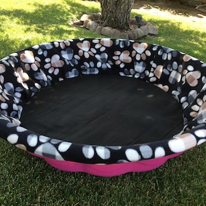 May include: A pink and black dog bed with a paw print and plaid pattern fleece lining. The bed is round and has a black fabric bottom.