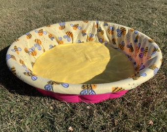 Peut inclure: Un lit pour animal domestique rose et jaune avec un motif d'abeilles. Le lit est fait d'un matériau doux et pelucheux et a un bord surélevé.