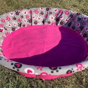 May include: Oval pet bed with a pink interior and a gray exterior. The gray exterior is patterned with pink hearts and black paw prints. The bed is sitting on green grass.