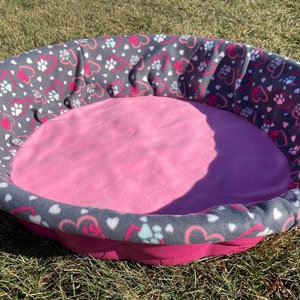 May include: A round pink and gray dog bed with a soft pink fleece lining and a gray and white fabric exterior with a pattern of hearts and paw prints.