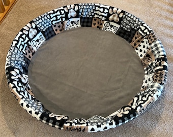 Peut inclure: Un lit pour chien rond avec un centre en tissu gris et une bordure en tissu patchwork noir et blanc. La bordure présente des empreintes de pattes, des os et le texte "Les chiens sont mes préférés".