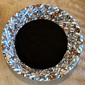 May include: Circular pet bed with a black center and a patterned border. The border features a blue and white gingham design with various dog illustrations in shades of brown, gray, and white. The bed is designed for comfort and style.