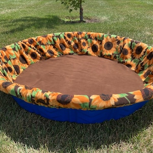 May include: A round pet bed with a blue base and a brown interior. The bed's rim is adorned with a sunflower pattern in shades of yellow, orange, and green. The bed is placed on a grassy surface.