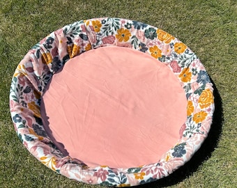 May include: A round, pink play mat with a floral print border. The border features a variety of flowers in shades of pink, yellow, and green.