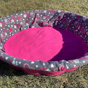 May include: A gray and pink dog bed with a heart and paw print design. The bed is made of soft fleece and has a raised rim.