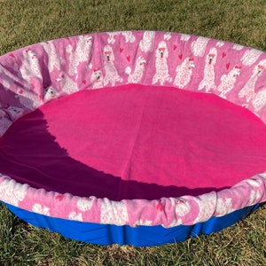 May include: Oval pet bed with a blue plastic base and a pink interior. The bed's rim is covered in a pink fabric patterned with white poodles wearing sunglasses and red hearts. The bed is designed for small pets.
