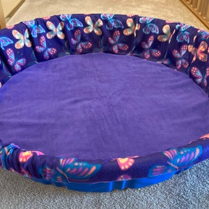 May include: A round pet bed with a blue base and a plush purple interior. The bed's rim is adorned with a fabric featuring a butterfly pattern in shades of blue, pink, and purple. This cozy bed is designed for small pets.