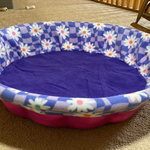 May include: A purple and white checkered pet bed with a pink rim and a daisy pattern. The bed is filled with a soft purple fabric.