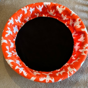 May include: A circular Halloween-themed pet bed with a black center and an orange rim. The rim is patterned with white ghost figures. The bed is designed for a small pet.