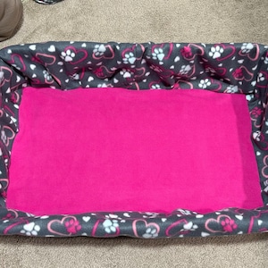May include: A gray and pink pet bed with a heart and paw print design. The bed has a pink fleece lining.