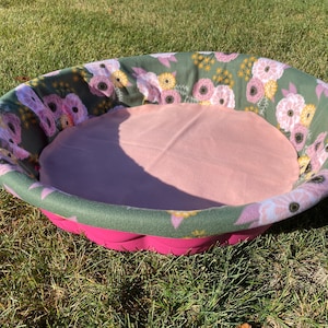 May include: A pink pet bed with a floral patterned rim and a soft, light pink interior. The rim features a green fabric with pink and yellow flower designs. The bed sits on a bright pink base, perfect for a small dog or cat.