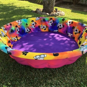 May include: A colorful dog bed with a purple fleece liner and a rainbow tie-dye print with paw prints and cartoon dogs. The bed is made of a soft, durable material and is perfect for small dogs.