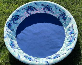 Peut inclure: Un lit rond pour animaux de compagnie avec un intérieur bleu et un extérieur à motifs. L'extérieur présente un fond bleu clair avec des illustrations de tortues marines bleues et turquoise. Le lit est conçu pour le confort et le soutien.