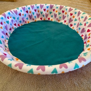 May include: A white pet bed with a teal blue fabric lining. The bed is decorated with a pattern of colorful hearts.