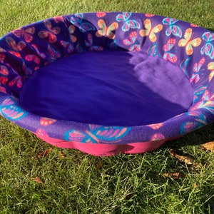 May include: A pink and purple pet bed with a butterfly print. The bed is made of soft fleece and has a comfortable, padded bottom.