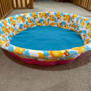 May include: A round pet bed with a pink base and a blue interior. The bed's rim is white, patterned with yellow rubber ducks and blue bubbles. The bed is designed for small pets.