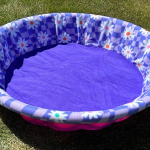 May include: Oval pet bed with a purple interior and a floral patterned rim. The rim features a checkerboard design with white daisies and orange centers. The bed has a pink base and is designed for small pets.