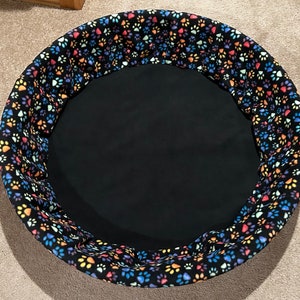 May include: A round pet bed with a black fabric interior and a colorful paw print patterned fabric exterior.