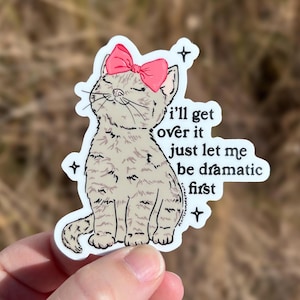 May include: A sticker featuring a cartoon cat with a pink bow, sitting and looking up. The text reads "I'll get over it just let me be dramatic first."
