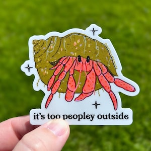 May include: A sticker featuring a cartoon hermit crab in a shell with the text "it's too peopley outside". The crab is red and the shell is brown with gold glitter. There are small gold stars around the crab.