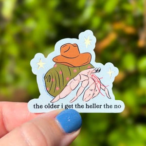 May include: A die-cut sticker featuring a light pink hermit crab wearing an orange cowboy hat, positioned on a light blue background with small yellow stars. The text "the older i get the heller the no" is printed in black at the bottom.