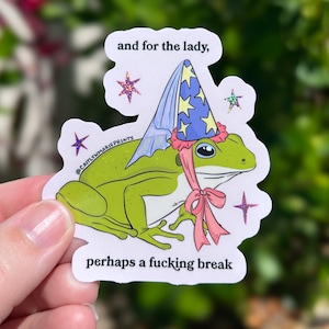 Pu&ograve; includere: Un adesivo raffigurante una rana verde con un cappello da mago con stelle blu e un nastro rosa. L'adesivo presenta il testo "and for the lady, perhaps a fucking break" e accenti di stelle. L'adesivo &egrave; tenuto da una mano.