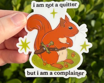 Può includere: Un adesivo fustellato raffigurante uno scoiattolo arancione che indossa una cintura marrone con una piccola spada. Il testo recita i am not a quitter in alto e but i am a complainer in basso. Stelle verde lime e ghiande sono sparse.