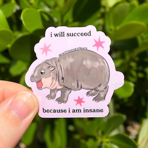 Puede incluir: Una pegatina troquelada con fondo rosa claro que presenta una ilustraci&oacute;n de un hipop&oacute;tamo gris. El texto i will succeed aparece en la parte superior y because i am insane en la inferior. Estrellas rosas rodean al hipop&oacute;tamo y el texto @CAITLYNMARIEPRINTS es visible sobre el lomo del animal.