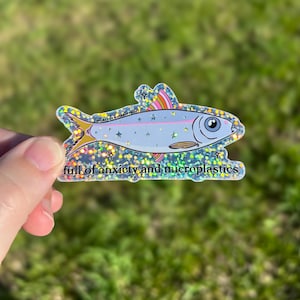 May include: A holographic sticker in the shape of a fish. The fish is light blue with a pink stripe and rainbow-colored fins. The text "full of anxiety and microplastics" is at the bottom of the sticker.